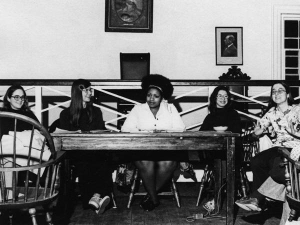Five women sit at wooden chairs behind a table. 