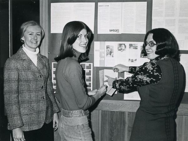 Three women smile in front of a display of materials about women and law 