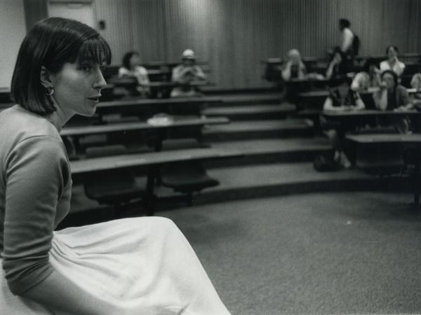 A woman sits in profile at the front of a classroom. Other people sit at desks in the background.