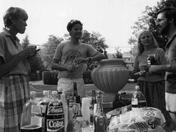 Four individuals stand around a table outside holding drinks. The table is covered with containers of chips and soft drinks. 